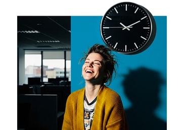 Smiling woman in a bright office space, standing against a bold blue backdrop with a wall clock, capturing the approachable, human side of Fleximize.